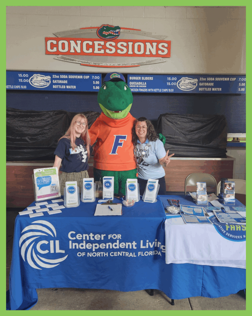 Two smiling CILNCF staff members pose on either side of the University of Florida mascot, Albert the Alligator, in front of a concession stand at Ben Hill Griffin Stadium. They’re standing behind a blue table covered with informational materials and brochures. The table prominently displays the logo and name: “Center for Independent Living of North Central Florida.” The scene is cheerful and welcoming, with the group making peace signs and showcasing community engagement at the Stomp the Swamp for Autism event.