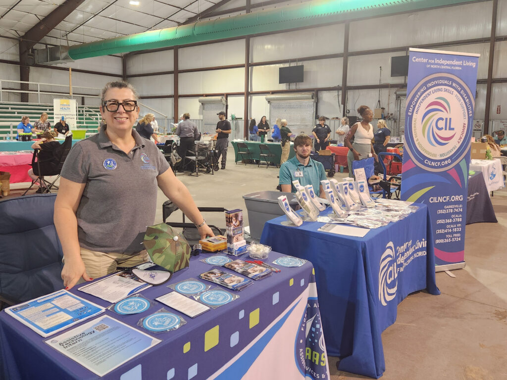 Claudia and Brent represent FAAST and the Center for Independent Living of North Central Florida at the 2nd Annual Disaster Preparedness and Community Resource Expo, held at the Suwannee River Fairgrounds. Claudia stands smiling behind the FAAST table, which is covered with brochures, stickers, and emergency planning materials. Brent sits at the adjacent CIL table, which displays informational flyers and promotional items in front of a large, branded banner. Other community organizations are visible in the background as attendees explore booths in the open-air indoor venue.