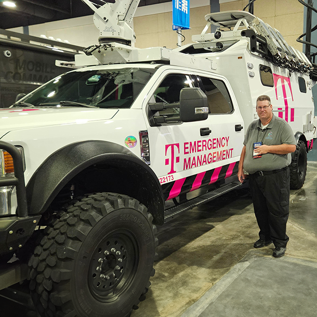 ID: Kevin stands next to a large white T-Mobile Emergency Management vehicle equipped with satellite and communication technology. The truck features bold pink branding and rugged off-road tires, indicating its use for disaster response. Behind the vehicle, other attendees and a Mobile Command Unit can be seen in the conference hall setting. Kevin is wearing a gray polo with the CIL logo and a conference badge, representing the Center for Independent Living of North Central Florida.