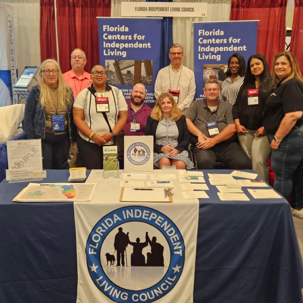 ID: A group of twelve individuals pose together at the Florida Centers for Independent Living booth during the Governor’s Hurricane Conference. They are standing and sitting in front of a display featuring banners that read “Florida Centers for Independent Living” and “Florida Independent Living Council.” The table in front of them is covered with brochures, informational materials, and a tablecloth with the Florida Independent Living Council logo. The setting is a conference exhibit hall with additional booths and banners visible in the background.