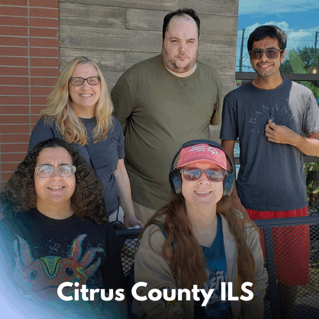 A group photo of five smiling people standing and sitting outside near a brick and wood wall. Two are seated in front, one wearing a colorful shirt and the other in a red cap, sunglasses, and headphones. Three stand behind them, smiling. Text overlay reads “Citrus County ILS.”