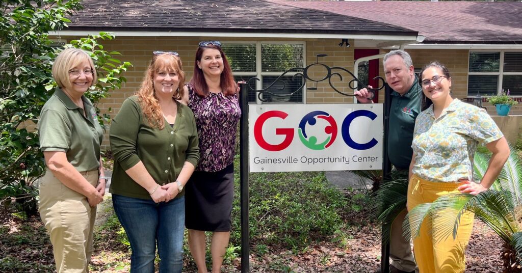 ID: Staff members from the CIL and Gainesville Opportunity Center smile together outside the GOC building next to a sign with the GOC logo.
