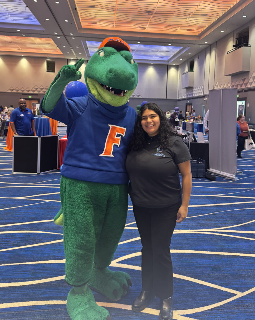 Jacee, Employment Consultant wearing a black polo shirt with the Center for Independent Living logo smiles while standing next to the University of Florida’s Albert the Alligator mascot. They are inside a large event space with blue carpeting, orange and blue UF décor, and people gathered in the background.