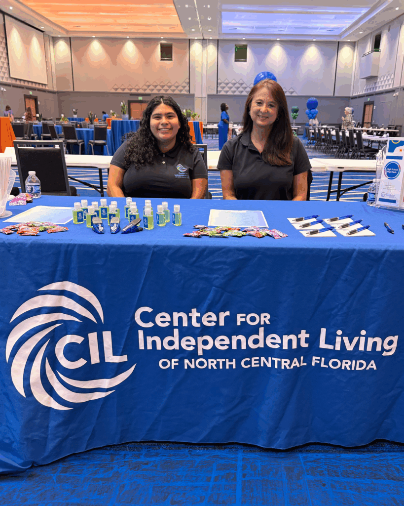Two women (Jacee and Mika) wearing black polo shirts with the Center for Independent Living logo sit behind a table covered with a blue CIL tablecloth. The table displays informational sheets, pens, hand sanitizers, and candy. The event is set up in a large room with blue carpeting, decorated tables, and balloon displays in the background.