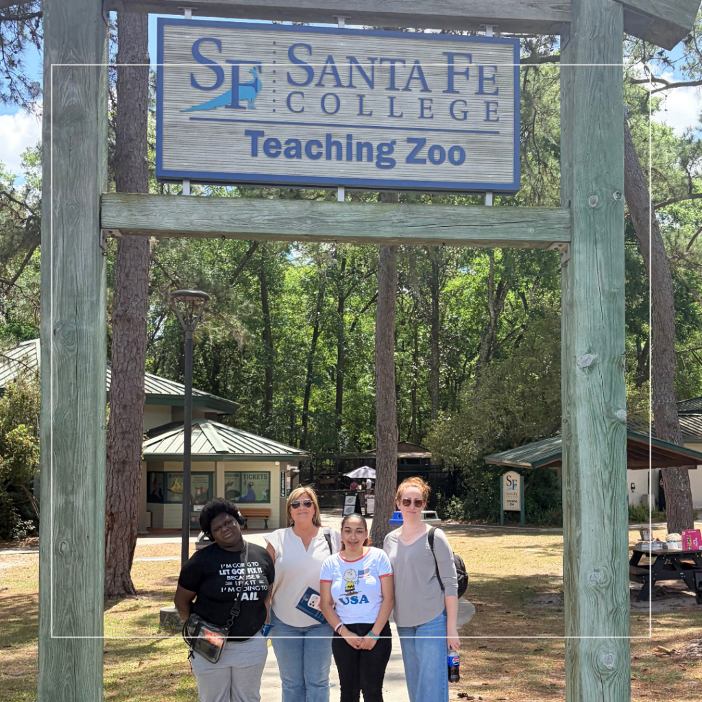 Four High School High Tech (HSHT) participants and a staff member pose for a group photo beneath the entrance sign for the Santa Fe College Teaching Zoo. The group stands outdoors on a sunny day, smiling in front of the zoo entrance, marking their career exploration visit focused on animal care, conservation, and postsecondary education opportunities. Trees and zoo facilities are visible in the background.