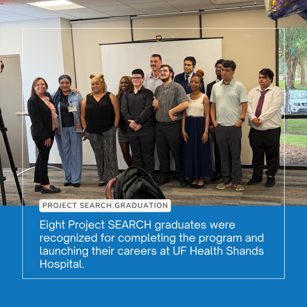 ID: A group of eight Project SEARCH graduates stand together with program staff, mentors, and supporters during a graduation celebration at UF Health Shands Hospital. The graduates are dressed in business attire and smiling as they pose for a group photo in front of a projection screen. A blue banner at the bottom of the image reads, "Project SEARCH Graduation," followed by the text, "Eight Project SEARCH graduates were recognized for completing the program and launching their careers at UF Health Shands Hospital." The event celebrates the graduates' successful completion of the internship program and their transition into competitive employment.