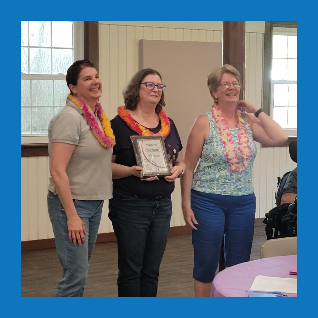 ID: Three CIL staff members (Amy, Andrea, Terri) wearing colorful leis stand together indoors at a celebration. The woman in the center holds a framed certificate recognizing Terri for her years of service. The women are smiling, with tables and other attendees partially visible around them.