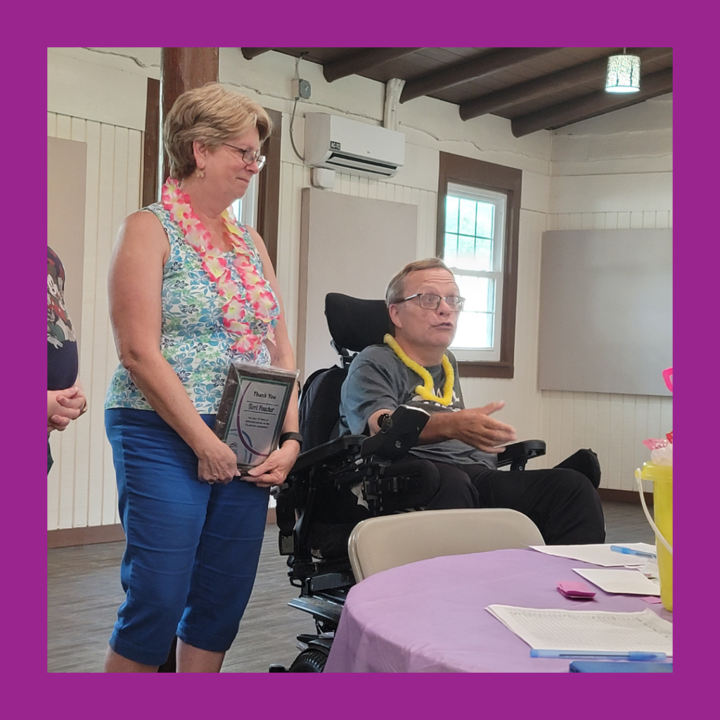 ID: Terri stands beside Mark seated in a power wheelchair during her retirement celebration. Both are wearing colorful leis, and Terri holds a framed certificate recognizing her years of service. The man gestures as he speaks while Terri listens beside him. A table with a purple tablecloth is visible in the foreground.